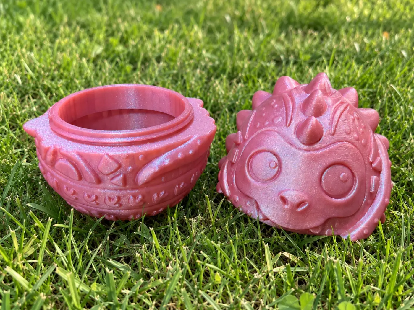 The two halves of a printed dragon egg on grass: the threaded bottom and the dragon-faced top